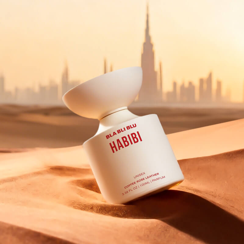 Perfume bottle labeled 'Habibi' by BLA BLU on sand with city skyline in the background