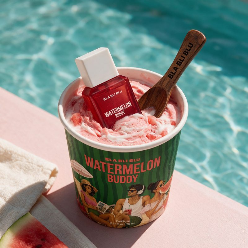 A cup of Watermelon Buddy frozen dessert sits by a swimming pool, with a matching red bottle and a wooden spoon placed inside the cup.