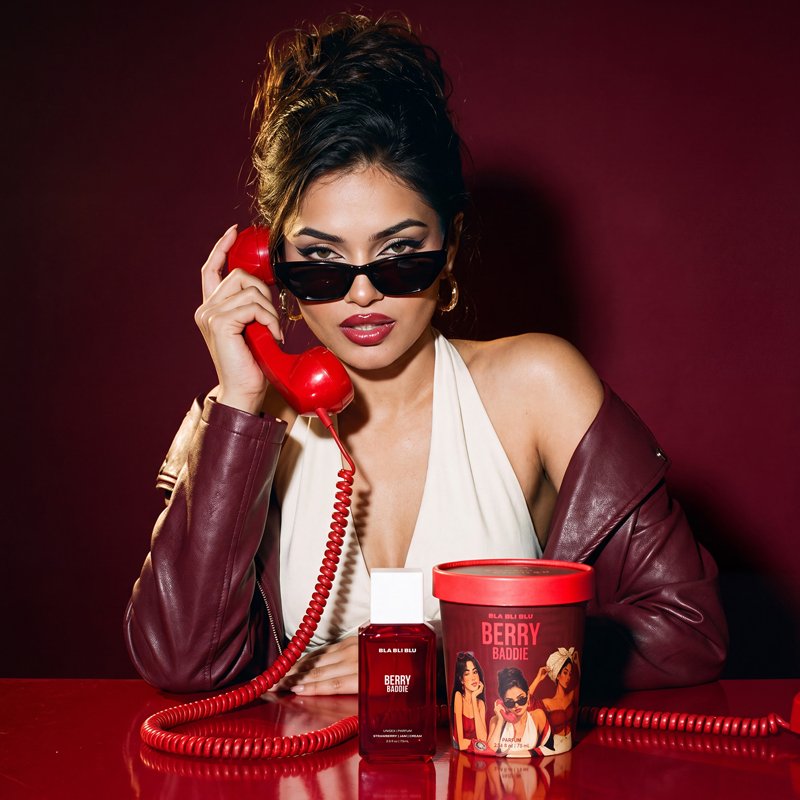 A woman in a white halter top and burgundy leather jacket sits at a red table, holding a red rotary telephone receiver. 