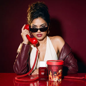 A woman in a white halter top and burgundy leather jacket sits at a red table, holding a red rotary telephone receiver. 