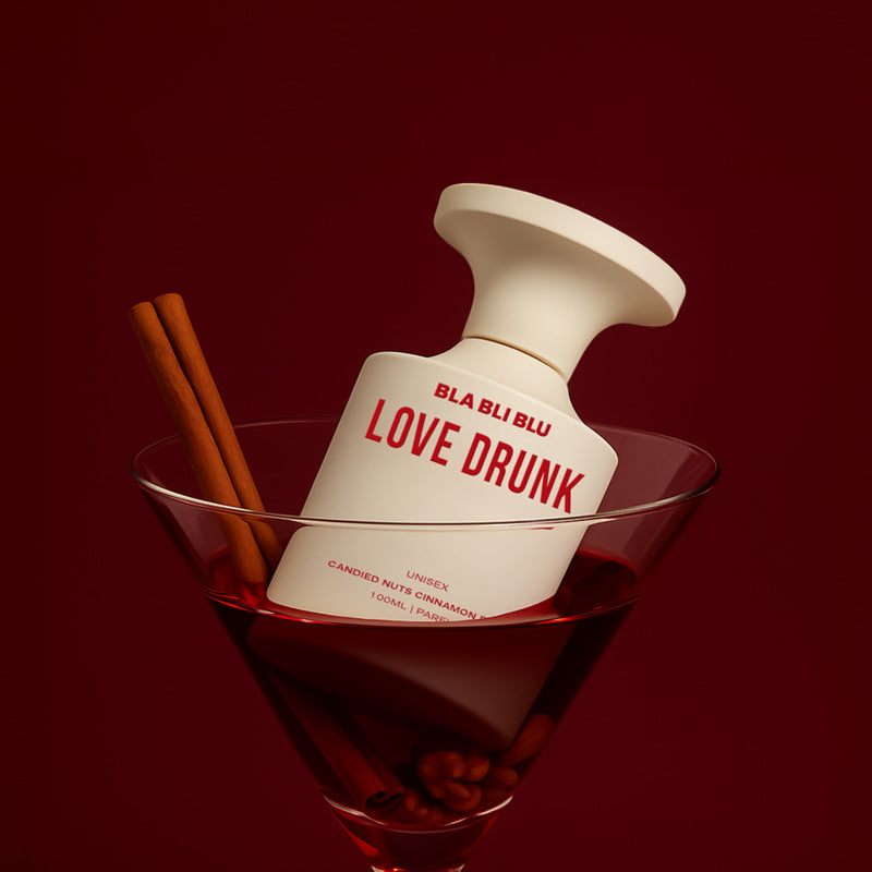 Perfume bottle labeled 'LOVE DRUNK' by BLA BLU in a glass with cinnamon sticks on a red background