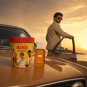 A man in a beige suit and sunglasses stands beside a vintage car at sunset, with two fragrance products in the foreground on the car hood: a yellow and red Mango So Coated tin.