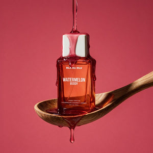 A small rectangular bottle labeled 'BLA BLI BLU WATERMELON BUDDY' filled with translucent pink-red liquid, resting on a wooden spoon.