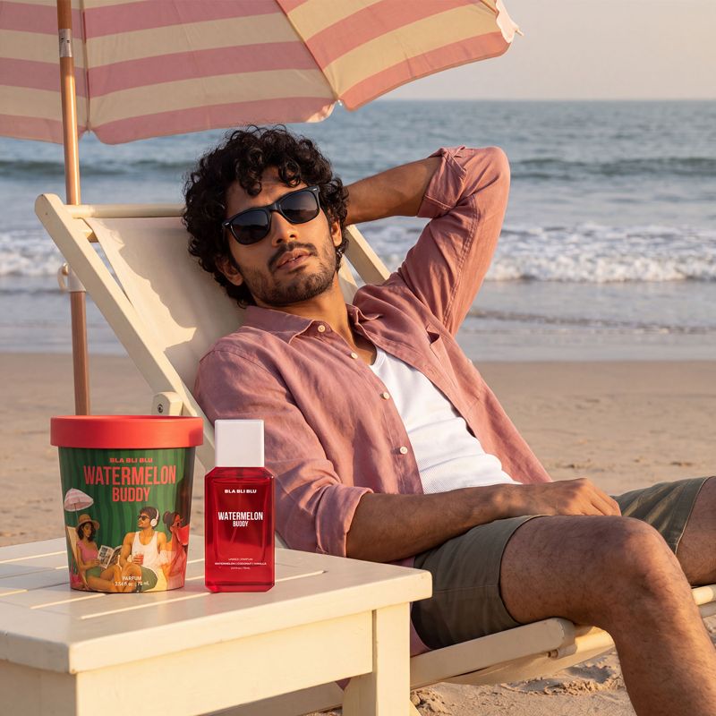 A man relaxes in a beach chair on the sand under a striped umbrella, with two bottles of Watermelon budy perfume.
